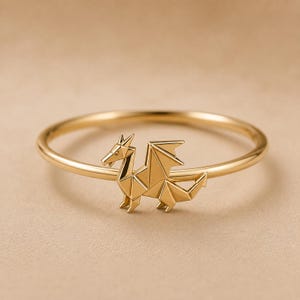 May include: A gold ring featuring a small, geometric dragon charm. The dragon is designed with sharp, angular lines that resemble an origami paper fold. The band of the ring is thin, smooth, and circular, maintaining a consistent gold tone throughout.