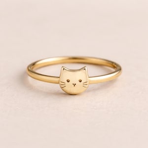 May include: A gold ring with a cat face design. The ring has a thin band and a detailed cat face with small eyes, a nose, and whiskers. The ring is a piece of jewellery.