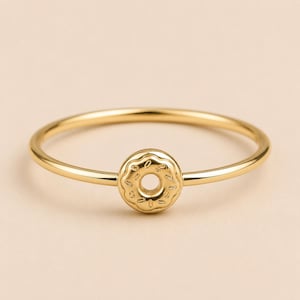 Op de afbeelding: Een goudkleurige ring met een kleine, ronde bedel in de vorm van een donut met reliëfspikkels op het oppervlak. De ringband is dun en glad en verbonden met het centrale donutmotief, dat een ronde opening in het midden heeft.