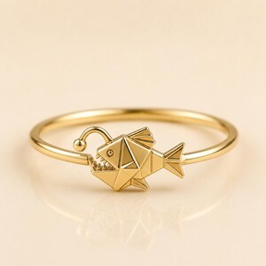 May include: A gold ring featuring a geometric anglerfish charm. The fish design includes a small, protruding lure on a curved stalk, sharp triangular teeth, and faceted body segments. The band is thin, smooth, and circular, crafted from polished gold.