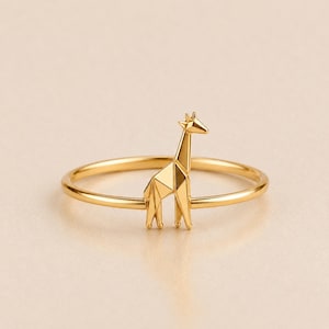 Puede incluir: Un anillo de tono dorado que presenta una figura de jirafa geométrica pulida. La jirafa está diseñada con facetas angulares nítidas que crean una apariencia estilizada inspirada en el origami. La banda es delgada, lisa y circular.