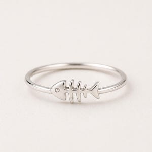 May include: A silver ring featuring a fish skeleton design. The ring has a thin band and a detailed fishbone motif, perfect for those who love aquatic themes. The ring is a simple yet elegant piece of jewelry.
