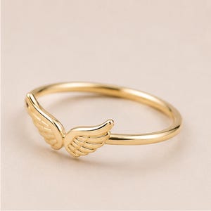K&ouml;nnte beinhalten: Ein goldfarbener Ring mit einem Paar detaillierter Fl&uuml;gel. Der Ring hat ein schmales Band, und die Fl&uuml;gel sind vorne positioniert, wodurch ein zartes und elegantes Design entsteht. Der Ring wird vor einem neutralen Hintergrund gezeigt.