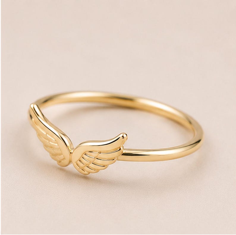 K&ouml;nnte beinhalten: Ein goldfarbener Ring mit einem Paar detaillierter Fl&uuml;gel. Der Ring hat ein schmales Band, und die Fl&uuml;gel sind vorne positioniert, wodurch ein zartes und elegantes Design entsteht. Der Ring wird vor einem neutralen Hintergrund gezeigt.