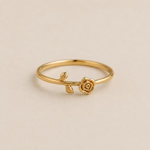 May include: A gold-toned ring featuring a thin band with a small, sculpted rose flower and two delicate leaves attached to the side. The metal has a polished, reflective finish, creating a smooth and continuous design along the circular band.