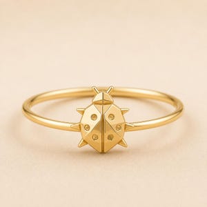 May include: A gold ring featuring a polished, geometric ladybug centerpiece. The ladybug has a faceted, angular design with small circular indentations on its wings and pointed protrusions along the sides, set on a thin, smooth, circular band.