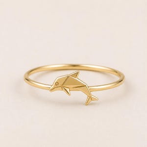 May include: A gold ring featuring a polished, geometric dolphin charm centered on a thin, circular band. The dolphin design is composed of angular, faceted planes that create a structured, low-profile silhouette on the delicate metal ring.