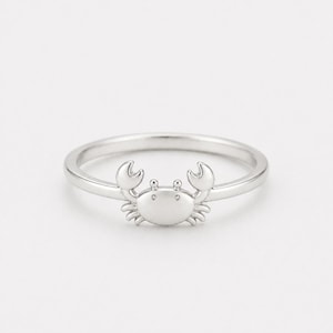 May include: A silver ring featuring a crab design. The crab has two claws and a rounded body with small details. The ring is made of a shiny, metallic material and is set against a plain background. The ring is a piece of jewelry.