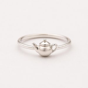 Peut inclure: Une bague argentée ornée d'un petit charme en forme de théière. La théière présente un design détaillé avec un bec, une anse et un couvercle. L'anneau de la bague est fin et poli, créant un bijou délicat et élégant. La bague est un accessoire charmant.