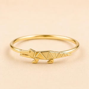 May include: A gold-toned ring featuring a polished finish and a geometric, stylised alligator or crocodile figure integrated into the band. The animal design is composed of sharp, angular lines and flat facets, creating a sleek, modern appearance.