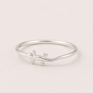 May include: A thin, polished silver-toned ring featuring a small, integrated lizard-shaped ornament along the band. The metal surface is smooth and reflective, with the lizard design seamlessly incorporated into the circular shape of the ring.