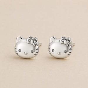 May include: A pair of silver-toned stud earrings shaped like the iconic Hello Kitty character. Each earring features the cat's face with its signature bow on the side, smooth metallic surfaces, and small engraved details for the eyes and nose.