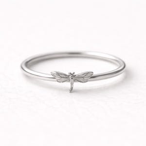 May include: A silver ring with a dragonfly motif. The ring has a slender band and a detailed dragonfly charm at the front. The dragonfly's wings and body are intricately designed. A delicate piece of jewellery, inspired by nature.