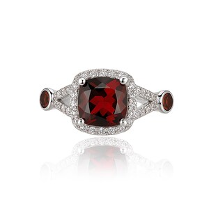 Peut inclure: Une bague en argent ornée d'une grosse pierre rouge foncé carrée, entourée de petites pierres claires. Deux petites pierres rouges rondes sont serties de chaque côté de la monture. La bague est présentée sur une surface blanche.