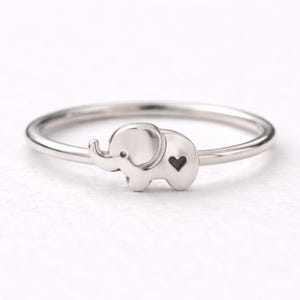 May include: A silver ring featuring a small elephant charm with a heart-shaped cutout. The ring has a thin band and a polished finish, suitable for everyday wear. The elephant charm is a cute and whimsical design.