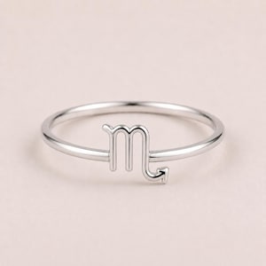 May include: A thin, polished silver-toned ring featuring a prominent Scorpio zodiac symbol as the centerpiece. The band is smooth and circular, with the astrological sign integrated directly into the design, showcasing a clean, metallic finish.
