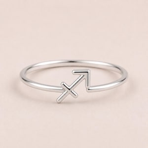 May include: A polished silver-toned ring featuring a Sagittarius zodiac symbol as the focal point. The band is thin and circular, with the arrow-shaped astrological sign integrated directly into the design, creating a continuous metallic loop.