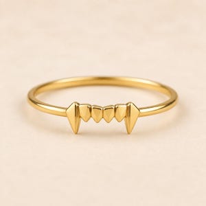 Peut inclure: Une fine bague en or présentant un motif central composé de six formes pointues en rangée, rappelant des dents. L'anneau est lisse et circulaire, avec l'élément décoratif intégré directement dans la partie supérieure de la structure.