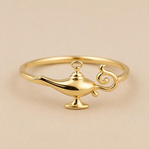 May include: A polished gold ring featuring a central charm shaped like a classic oil lamp. The lamp design includes a curved spout, a small handle, and a decorative base, all integrated into the thin, circular band of the ring with a smooth finish.
