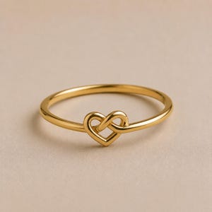 May include: A polished gold-tone ring featuring a central heart-shaped motif. The band is thin and circular, with the metal looping to form a continuous, stylised heart shape at the front, creating a smooth, reflective surface throughout the piece.