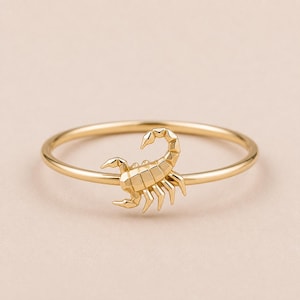 Puede incluir: Un anillo fino de oro con un pequeño dije de escorpión facetado centrado en la banda. El escorpión se muestra con la cola curvada hacia arriba y las patas extendidas, exhibiendo un acabado metálico pulido y detalles geométricos angulares.