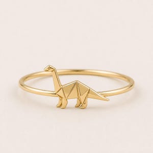 May include: A gold-toned ring featuring a thin, circular band and a central ornament shaped like a long-necked dinosaur. The dinosaur figure is designed with geometric, angular lines that mimic the appearance of folded paper origami.