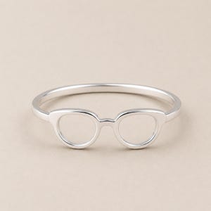 Op de afbeelding: Een ring van gepolijst zilver met een centraal ontwerp in de vorm van een bril. De band is glad en cirkelvormig en sluit aan op het montuur van de bril, dat bestaat uit twee open, afgeronde randen die verbonden zijn door een kleine brug.