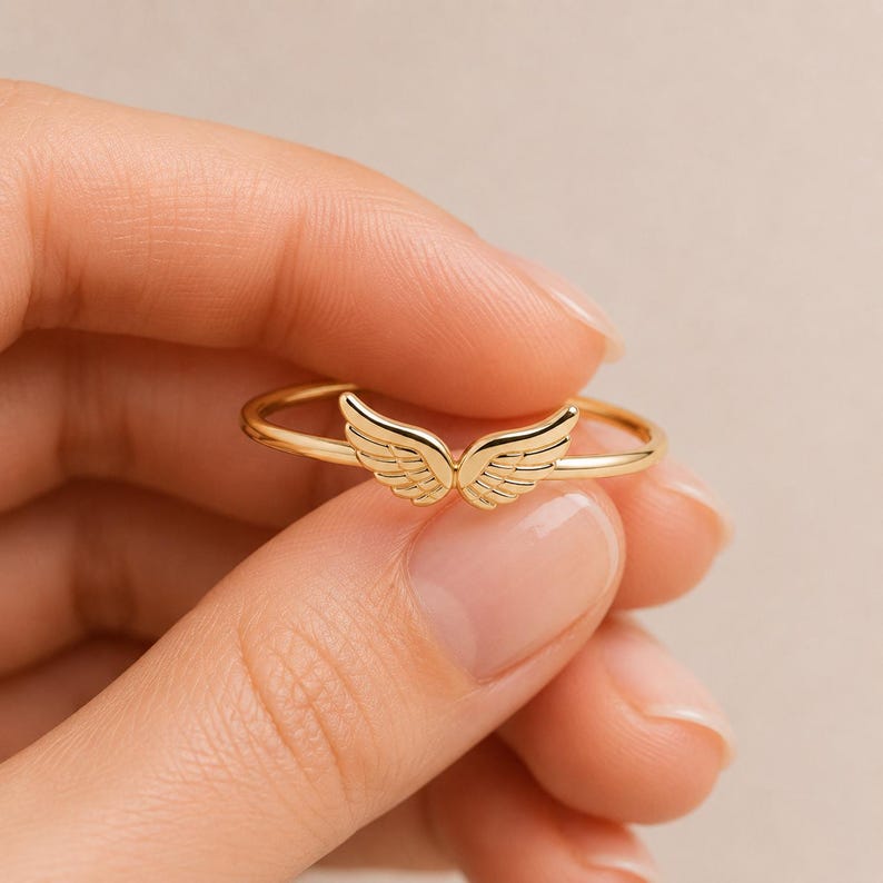 K&ouml;nnte beinhalten: Ein goldfarbener Ring mit einem filigranen Fl&uuml;geldesign. Die Fl&uuml;gel sind mit feinen Linien versehen, und der Ring ist glatt und poliert. Der Ring wird in einer Hand vor einem weichen, neutralen Hintergrund gehalten. Dieser Ring ist ein Schmuckst&uuml;ck.