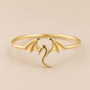May include: A gold-toned ring featuring a central dragon motif with outstretched wings. The dragon design is integrated into the thin, circular band, creating a continuous silhouette. The metal has a smooth, polished finish throughout the piece.