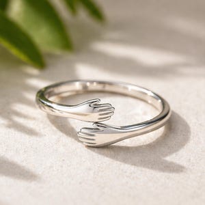 Puede incluir: Un anillo plateado con un diseño de dos manos abrazándose. El anillo está hecho de un metal brillante y tiene un acabado liso y pulido. Las manos están detalladas con dedos y están posicionadas para que parezca que se están abrazando.