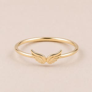 K&ouml;nnte beinhalten: Ein goldfarbener Ring mit einem Fl&uuml;geldesign. Der Ring hat ein d&uuml;nnes Band und ein Paar detaillierte Fl&uuml;gel in der Mitte. Die Fl&uuml;gel sind symmetrisch und haben eine strukturierte Oberfl&auml;che. Der Ring ist vor einem neutralen Hintergrund platziert.