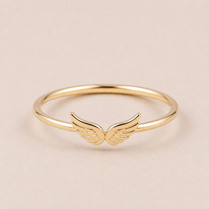 K&ouml;nnte beinhalten: Ein goldfarbener Ring mit einem Fl&uuml;geldesign. Der Ring hat ein d&uuml;nnes Band und ein Paar detaillierte Fl&uuml;gel in der Mitte. Die Fl&uuml;gel sind symmetrisch und haben eine strukturierte Oberfl&auml;che. Der Ring ist vor einem neutralen Hintergrund platziert.