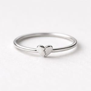 May include: A thin, polished silver-tone metal ring featuring a small, centered heart charm. The heart charm is divided down the middle by a jagged, lightning-bolt-shaped line, creating the appearance of a broken heart design on the band.