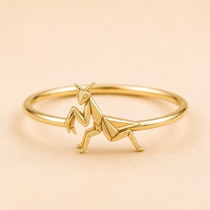 May include: A gold ring featuring a polished, geometric, origami-style praying mantis ornament centred on a thin, circular band. The insect design is composed of sharp, angular facets that create a structured, three-dimensional appearance.