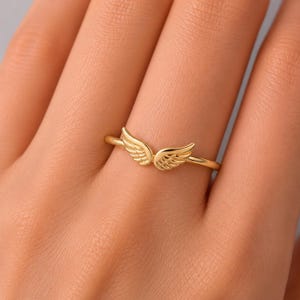 K&ouml;nnte beinhalten: Ein goldfarbener Ring mit einem Paar detaillierter Engelsfl&uuml;gel. Die Fl&uuml;gel sind so positioniert, dass sie sich um den Finger kr&uuml;mmen und ein zartes und elegantes Design bilden. Der Ring wird an einer Hand gezeigt.