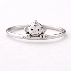 May include: A silver-colored ring featuring a small, smiling monkey design. The monkey has detailed features, including ears, eyes, and hands, and is positioned on the ring's band. The ring is a delicate piece of jewelry.
