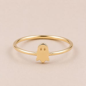 May include: A gold-colored ring featuring a small ghost-shaped charm. The ring has a thin band and a simple, minimalist design. The ghost charm has two small eye details.