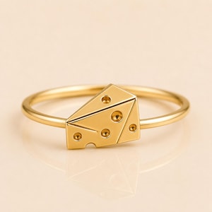 Puede incluir: Un anillo de tono dorado que presenta un pequeño dije triangular con forma de cuña de queso suizo. El dije tiene una superficie lisa y pulida con varias hendiduras circulares de diferentes tamaños, imitando la apariencia de los agujeros.