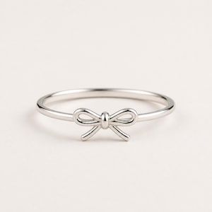 May include: A silver-colored ring featuring a delicate bow design. The bow is centered on the band, creating a simple yet elegant look. The ring is likely made of sterling silver or a similar metal, and is suitable for everyday wear.