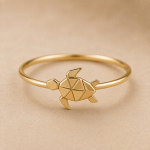 Peut inclure: Une bague dorée ornée d'un motif central en forme de tortue géométrique. Le design de la tortue est composé de multiples facettes triangulaires, créant une apparence structurée et angulaire. L'anneau est fin, lisse et circulaire, de couleur dorée.