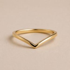 May include: A polished gold ring featuring a distinct V-shaped chevron design. The band has a smooth, reflective surface with a uniform metallic finish, creating a sharp point at the centre of the ring that tapers towards the back of the band.