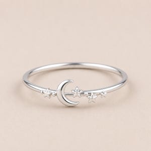 Könnte beinhalten: Ein dünner silberfarbener Ring mit einem polierten Mondsichel-Element in der Mitte des Bandes, flankiert von vier kleinen sternförmigen Akzenten in unterschiedlichen Größen. Das Metall hat ein glattes, reflektierendes Finish.