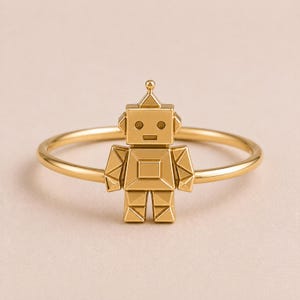 Può includere: Un anello color oro con un piccolo ciondolo a forma di robot geometrico centrato su una sottile fascia circolare. Il robot ha una testa quadrata con un'antenna appuntita, occhi circolari, una bocca rettangolare e un corpo segmentato.