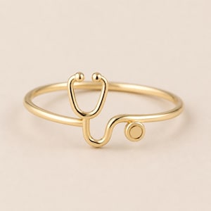 Puede incluir: Un anillo de oro pulido que presenta un diseño delicado y estilizado de estetoscopio integrado en la banda. El metal es liso y reflectante, mostrando la forma icónica del instrumento médico con una pieza torácica circular y tubos.