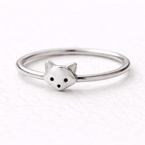 May include: A silver-colored ring featuring a fox head design. The fox head is the focal point, with small black dots for eyes and a tiny nose. The ring band is thin and smooth, creating a minimalist aesthetic.
