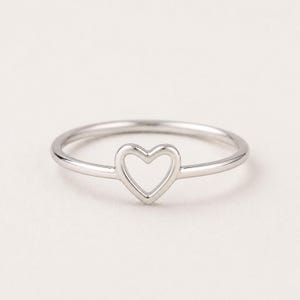 May include: A silver ring with a heart-shaped cutout. The ring is thin and delicate, with a polished finish. The heart is centered on the band, creating a simple yet elegant design. This ring is a piece of jewelry.