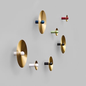 May include: A collection of modern wall hooks with a brass finish and colorful accents. The hooks feature a circular design with a brass disc and a cylindrical post in various colors, including white, black, blue, red, and green. The hooks are mounted on a gray wall.
