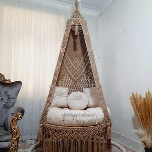 May include: A beige macrame hanging chair with a round seat and backrest, adorned with tassels and pillows. The chair is suspended from the ceiling and features the text "KNOTS&HANGINGS".