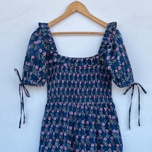 May include: A navy blue dress with a floral pattern. The dress has a square neckline, short sleeves with tie details, and a fitted bodice that flares into a flowing skirt. The dress is hanging on a wooden hanger.