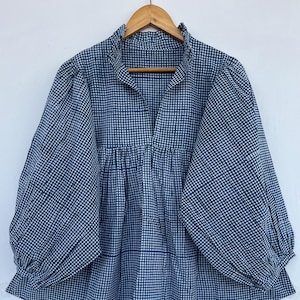 Puede incluir: Una blusa a cuadros azul y blanco con mangas largas y voluminosas y un cuello con volantes. La blusa está colgada de una percha de madera sobre un fondo blanco. La tela tiene un pequeño patrón cuadrado.